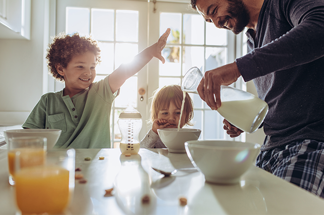 Dad pouring milk into a bowl and two kids sitting at the table having breakfast